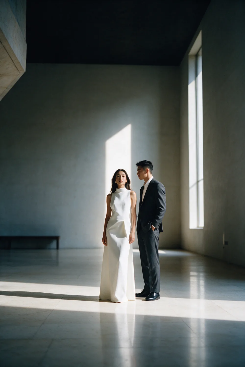 A couple in a pale hall crossed by a single slab of window light.